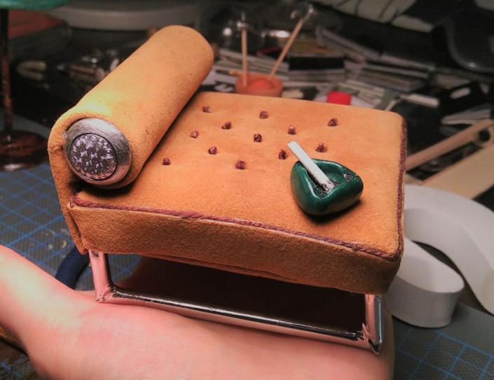 Recamiére und Aschenbecher, 1:12 Miniatur einer Recamière und eines Aschenbechers im Maßstab 1:12, Recamiere aus hellbraunem Wildleder mit einem silbernen Gestell, Aschenbecher aus grünem Kunststoff mit einer glimmenden Zigarette, auf einer Handinnenfläche stehend, im Hintergrund Werkzeug und Material