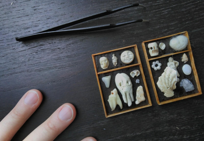 Miniaturen, zwei Holzrahmen aus hellem Holz mit dunklem Hintergrund, darin verschiedene kleine Elemente wie Zähne, Muscheln, Gesichter, Perlen, Steine in verschiedenen Weiß- und Naturtönen