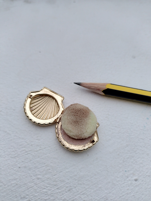 Miniatur-Puderdose in Form einer goldenen Muschel, abgebildet mit Bleistift für Größenvergleich