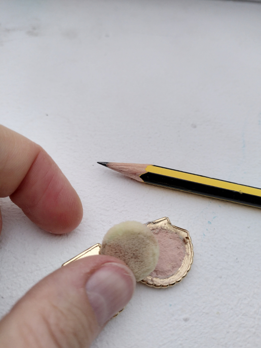 Miniatur-Puderdose in Form einer goldenen Muschel, abgebildet mit Bleistift für Größenvergleich