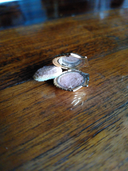 Puderdose zum Sturmgrünen Waschraum, sie hat die Form einer Muschel und liegt mit einem sehr kleinen Puderschwamm auf einem Holzuntergrund