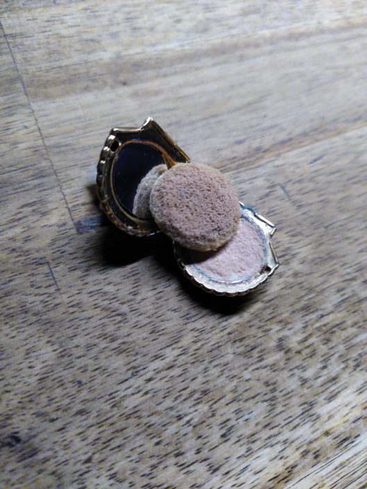 Puderdose zum Sturmgrünen Waschraum, sie hat die Form einer Muschel und liegt mit einem sehr kleinen Puderschwamm auf einem Holzuntergrund