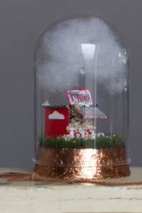 Foto einer Miniatur: eine rot-weiße Popcorn-Bude, die auf einer Wiese unter einer weißen Wolke steht, geschützt von einer Glaskuppel, auf einem rotgoldenem Sockel