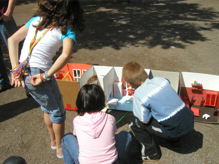 Drei Kinder im Grundschulalter schauen eine aus Pappe gebaute Puppenwohnung an