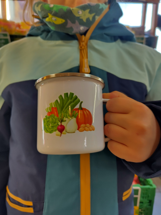 Emailletasse mit der Illustration vom Gemüse aus dem Logo des Gemüseladens und einem dekorativen Radieschen gehalten von einer Kinderhand, vor einer Jacke in Blautönen