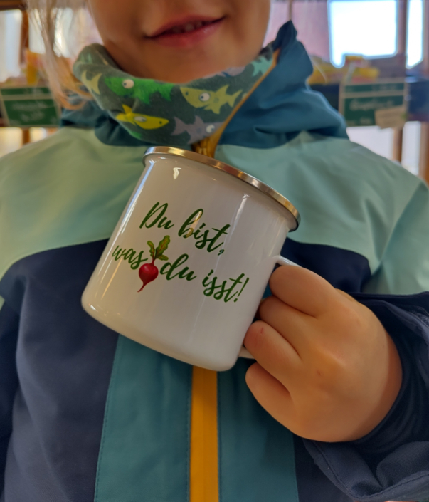 Emailletasse mit der Aufschrift "Du bist was du isst" und einem dekorativen Radieschen gehalten von einer Kinderhand, vor einer Jacke in Blautönen, Gesicht im Anschnitt, nur ein lächelnder Mund