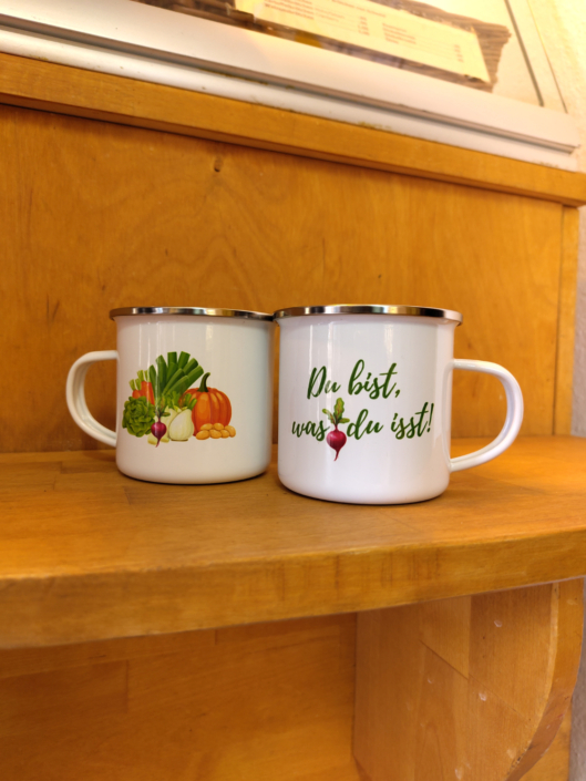 zwei Emailletasse auf einer Holztheke stehend, eine mit dem Gemüse aus dem Logo und eine mit der Aufschrift "Du bist was du isst!"