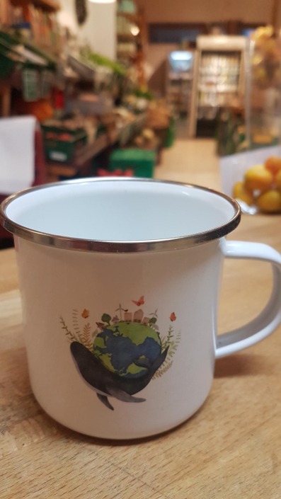 Tasse aus weißen Emaille bedruckt mit einer Illustration eines Wals, der die Welt trägt auf der Theke im Gemüseladen