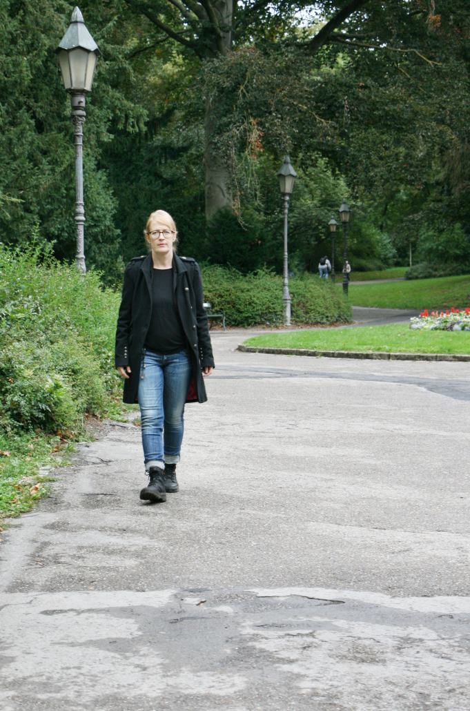 Rike Freeman geht durch den Würzburger Ringpark, sie trägt Boots, Jeans, ein schwarzes Oberteil und einen schwarzen Mantel, sie geht auf den:die Betrachtenden zu